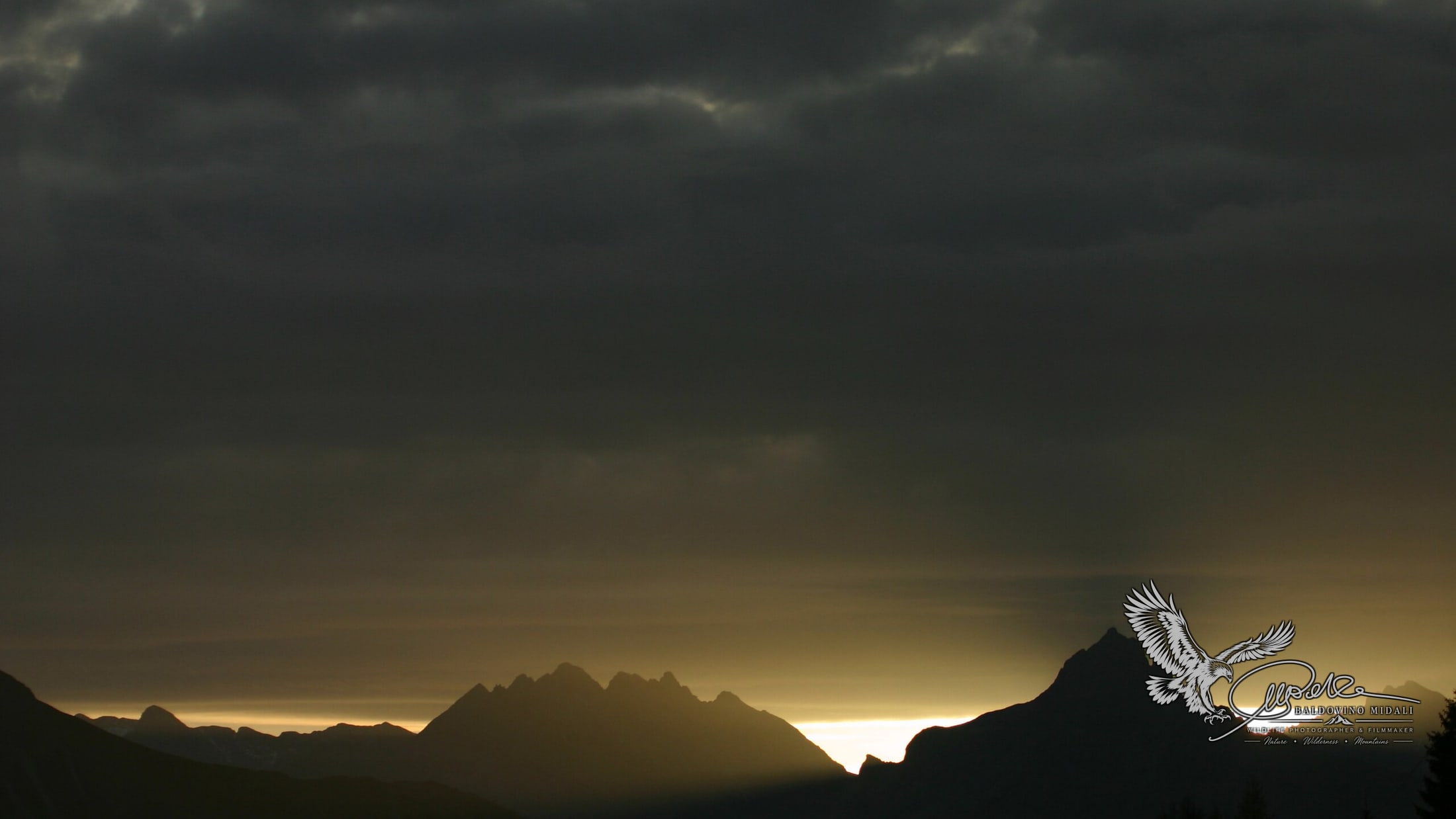 Profilo delle montagne al tramonto sotto un cielo scuro e nuvoloso, con luce radente sull'orizzonte