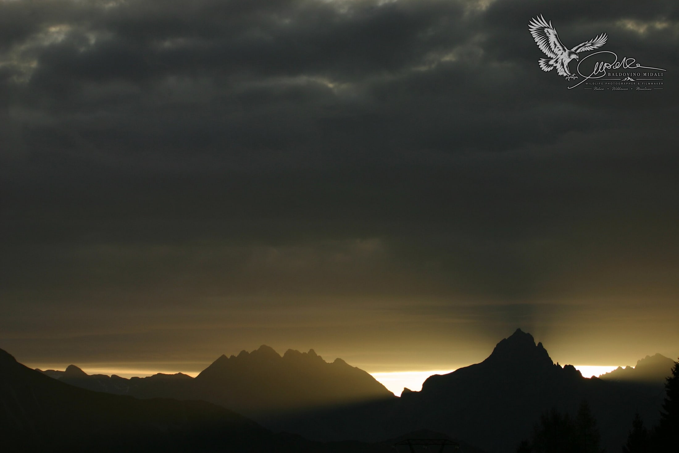 Profilo delle montagne al tramonto sotto un cielo scuro e nuvoloso, con luce radente sull'orizzonte